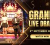 Hosts stand on stage during the Jumbo Ticket Grand Live Draw event, with a large jackpot machine and a bright crown backdrop behind them.